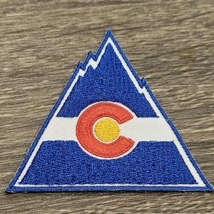 May include: A triangular patch featuring the Colorado state logo. The patch has a blue background with a white border and a white stripe across the centre. The logo includes a red "C" with a yellow circle inside, and white mountain peaks at the top.