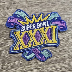 Puede incluir: Un parche bordado azul, morado y dorado con las palabras "Super Bowl XXXI" en un diseño de corona.