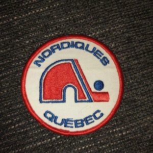 May include: A white, red, and blue embroidered patch with the words "Nordiques Quebec" and a stylized hockey stick and puck.