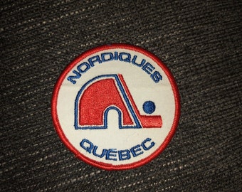 May include: A white, red, and blue embroidered patch with the words "Nordiques Quebec" and a stylized hockey stick and puck.