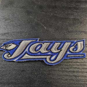 May include: Embroidered patch with the word "Jays" in a stylized font, featuring a blue bird head design on the left. The patch is blue, grey, and white, with black outlining.