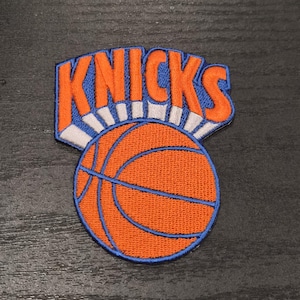 May include: An embroidered fabric patch featuring the word KNICKS in bold orange letters with a blue outline, positioned above a round orange basketball graphic with blue lines. The design is finished with a blue border and white accents.
