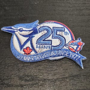 Può includere: Toppa ricamata con logo di ghiandaia azzurra, la scritta "25th Season" e "Summer Tradition Since 1977". La toppa ha sfondo bianco con dettagli blu e rossi, e una piccola foglia d'acero.