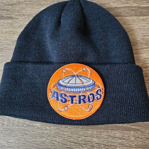 May include: Navy blue knitted beanie hat with a circular patch. The patch features the word "ASTROS" in white lettering, set against an orange background with a blue and white stadium design. A classic winter hat.
