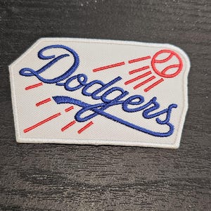Può includere: Una toppa bianca con la scritta "Dodgers" in blu, accompagnata da una palla da baseball e linee rosse. La toppa ha un bordo bianco, probabilmente un emblema di una squadra di baseball.