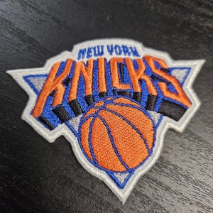 Op de afbeelding: Geborduurde patch met het logo van de New York Knicks. De patch heeft een witte rand met de woorden "New York" boven "Knicks" in oranje en blauw. Een oranje basketbal staat in het midden.