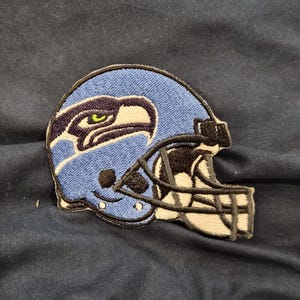 May include: Embroidered patch of a blue football helmet with a black facemask. The helmet features a white and navy blue logo of a bird with a green eye. The patch is on a dark blue fabric.