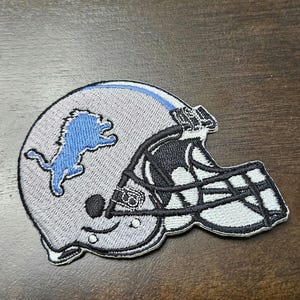 May include: An embroidered patch depicting a football helmet with a lion mascot. The helmet is white with a black facemask and a blue lion with a white outline.