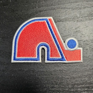 Peut inclure: Un écusson brodé rouge, blanc et bleu avec une lettre "A" stylisée et une rondelle de hockey. L'écusson est sur un fond de bois foncé.