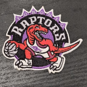 May include: Embroidered patch featuring the Toronto Raptors logo. The patch shows a red raptor holding a basketball, set against a black and purple background. The word "RAPTORS" is arched above the raptor in white letters.