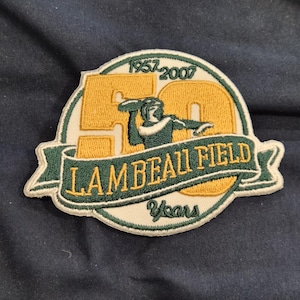 以下が含まれることがあります： 緑と金のデザインの刺繍パッチ。「LAMBEAU FIELD」の文字がバナーに、数字の「50」、年号の「1957-2007」が描かれています。アメフトのボールを投げる姿も描かれています。