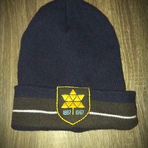 Puede incluir: Un gorro de punto azul marino con una franja marrón y blanca y una hoja de arce bordada en amarillo con los años 1867 y 1967.