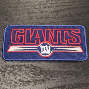 May include: A rectangular embroidered patch with a dark blue background. The word GIANTS is stitched in red with a blue outline. Below the text is a white horizontal bar with a central circle containing the lowercase letters ny in white.