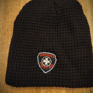 May include: Black knitted beanie with a red and white embroidered patch that says "Emergency First Aid".