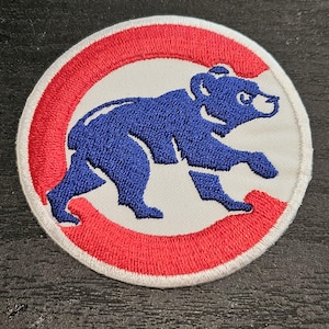 Puede incluir: Parche bordado con el logotipo de los Chicago Cubs. El parche presenta un oso azul dentro de un círculo rojo sobre un fondo blanco. El oso está representado en movimiento.