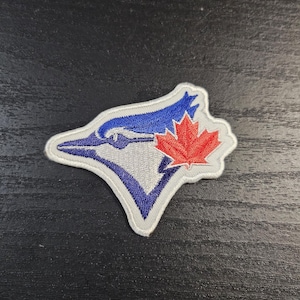 May include: Embroidered patch featuring the Toronto Blue Jays logo. The logo shows a stylized blue jay head with a white and blue design, and a red maple leaf. The patch has a white border and is likely for apparel.