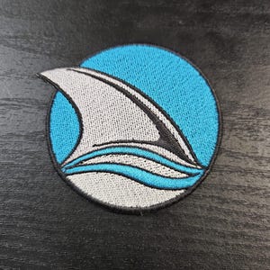 May include: Embroidered patch featuring a shark fin design. The fin is white with black accents, set against a light blue circle. Below the fin are two white and blue wavy lines, all outlined in black.