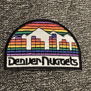 May include: A colorful embroidered patch with a rainbow mountain skyline and the text "Denver Nuggets".