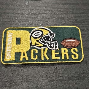 Könnte beinhalten: Gestickter Green Bay Packers Patch mit einem gelben "P" und den Worten "GREEN BAY" und "PACKERS" in weißer und gelber Schrift. Ein gelber Footballhelm und ein brauner Football sind ebenfalls auf dem Patch gestickt.