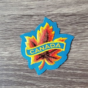 Puede incluir: Parche bordado con una hoja de arce colorida en tonos rojos, naranjas y amarillos, sobre un fondo azul. La palabra "CANADA" está bordada en amarillo. Ideal para chaquetas, bolsos u otros textiles.