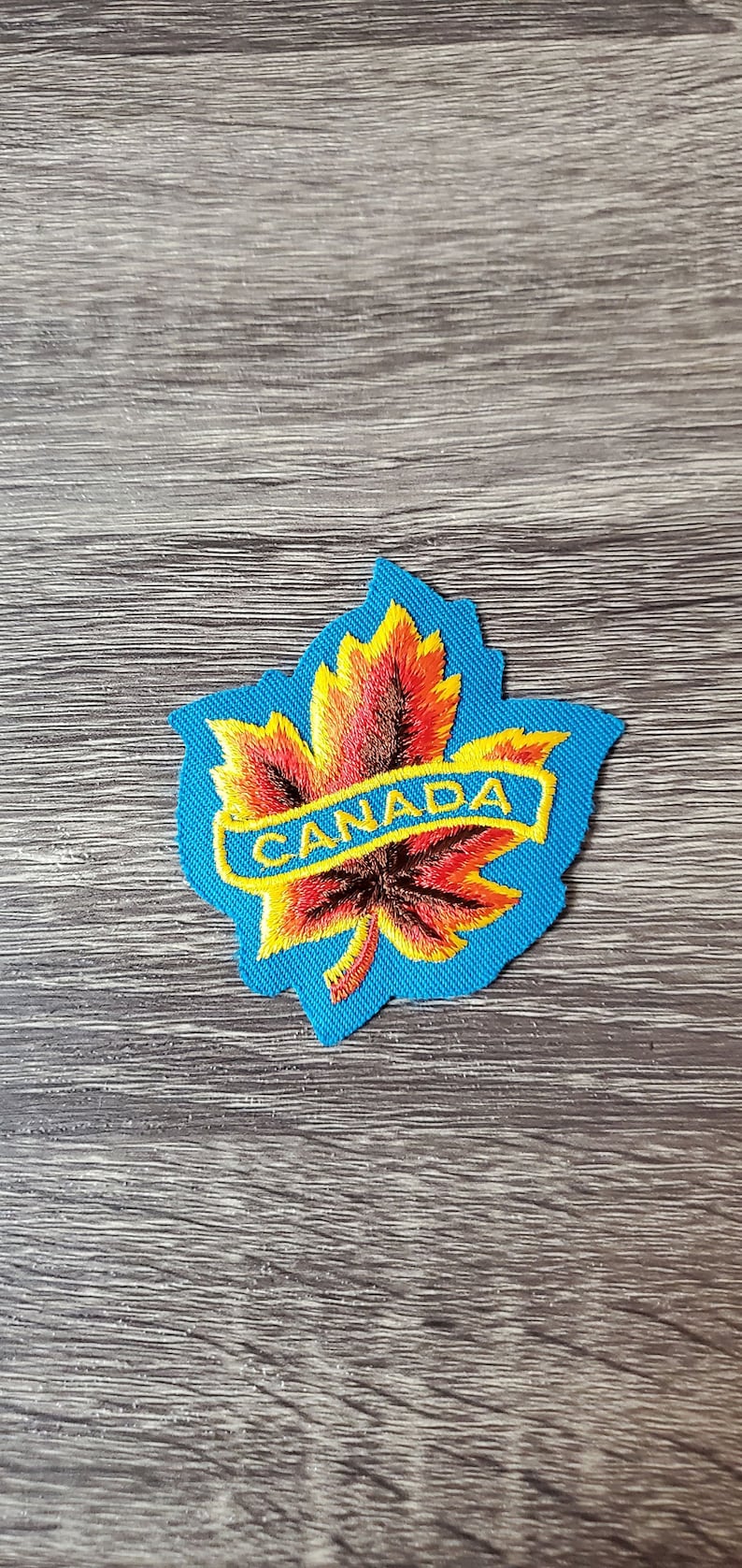 May include: Embroidered patch depicting a maple leaf in shades of red, orange, and yellow, on a blue backing. The word "CANADA" is embroidered in yellow. Suitable for attaching to clothing or accessories.