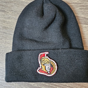 Puede incluir: Gorro de punto negro con borde doblado. El gorro presenta un parche con un logotipo rojo, dorado y blanco de una figura con casco. El gorro está hecho de un material de punto acanalado suave.