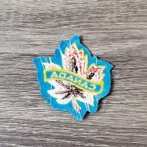 May include: A blue, leaf-shaped patch with the word "CANADA" in yellow letters. The leaf design features red, white, and brown details, set against a blue border. The patch is placed on a wooden surface.