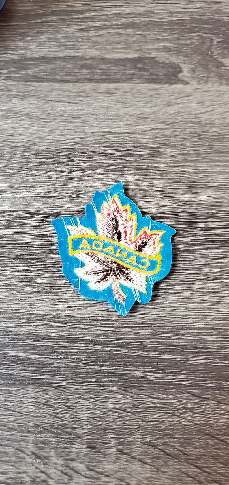 May include: A blue, leaf-shaped patch with the word "CANADA" in yellow letters. The leaf design features red, white, and brown details, set against a blue border. The patch is placed on a wooden surface.