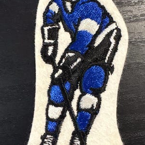 Puede incluir: Parche bordado que representa a un jugador de hockey en acción, en azul, blanco y negro, con un palo de hockey. El parche, sobre fondo blanco, está diseñado para coserse en ropa o accesorios. Tema deportivo.