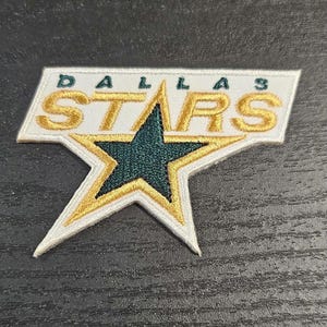 Puede incluir: Parche bordado con el logotipo de los Dallas Stars. El parche tiene un fondo blanco con las palabras "DALLAS STARS" en letras doradas. Una estrella verde con ribete dorado está en el centro. El parche es rectangular con forma de estrella.