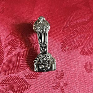 Peut inclure: Épinglette en métal argenté en forme de coupe de trophée avec des détails complexes. Le trophée a une base ronde et un corps haut et mince avec un sommet décoratif.