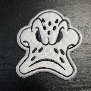 May include: Embroidered patch of a cartoon ghost-like figure with a sad expression. The patch is white with black details, including eyes, spots, and a curved mouth. The patch has a black border.