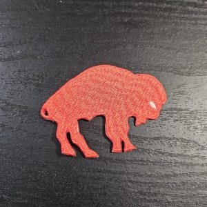 May include: A red embroidered patch in the shape of a buffalo. The patch has a white accent on the buffalo's head and is set against a dark background. The buffalo is facing right.