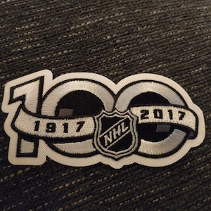 Puede incluir: Parche bordado que conmemora el centenario de la NHL. El parche presenta el número "100" en un diseño estilizado, con "1917" y "2017" en una cinta. El logotipo de la NHL está centrado. El parche es principalmente negro, blanco y gris.