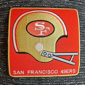 May include: A red and gold embroidered patch featuring a football helmet with the letters "SF" inside. The text "SAN FRANCISCO 49ERS" is below the helmet.