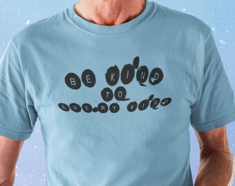 May include: A man wearing a light blue t-shirt with the text "Be Kind to Every Kind" in black lettering. The text is in a circle design. He is holding a book in his right hand.