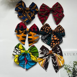 May include: Six fabric hair bows with diverse patterns and colours. The bows display vibrant African wax print designs in shades of blue, red, yellow, green, and brown. Each bow is tied in a classic bow shape.