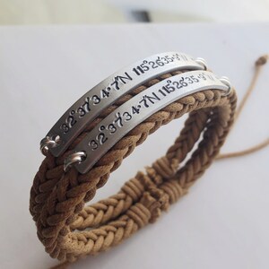 May include: A brown braided cord bracelet with two silver metal plates engraved with the coordinates "32°37′34.7″N 115°2635.9"