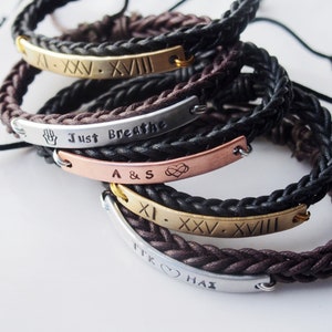 May include: A stack of five braided leather bracelets with metal plates engraved with different messages. The top bracelet has Roman numerals, the second says "Just Breathe", the third says "A & S" with a heart symbol, the fourth has Roman numerals, and the bottom bracelet says "TTK ♥  HAI".