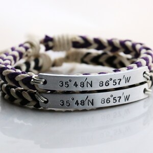 May include: Two braided bracelets with purple and white cords and silver metal tags engraved with the coordinates "35° 48'N 86° 57'W".