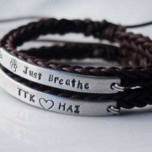 May include: Two brown braided bracelets with silver metal plates. The top plate has the text "Just Breathe" and a hamsa hand symbol. The bottom plate has the text "TTK ♥ HAI".