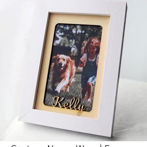 May include: White wooden picture frame with a photo of a girl and a golden retriever dog. The name "Kelly" is laser cut in gold script. The frame has a light yellow mat.