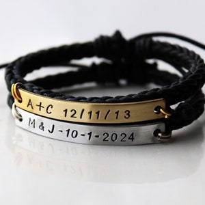 May include: Two personalized bracelets with black braided cords. The top bracelet has a gold metal band with the text "A+C 12/11/13". The bottom bracelet has a silver metal band with the text "M&J -10-1-2024".