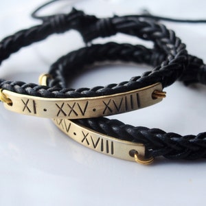 May include: Two black braided leather bracelets with gold-colored rectangular nameplates. The nameplates have Roman numerals engraved on them. The bracelets are on a white surface.