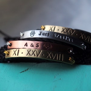May include: Four stacked bracelets with different engraved messages. The top bracelet is silver and says "Just Breathe" with Roman numerals. The second bracelet is copper and says "A & S" with a heart symbol. The third bracelet is gold and says "XI. XXV. XVIII". The bracelets are on a black cord.