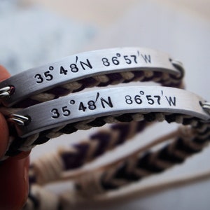 May include: Two silver bracelets with black and white braided cords. The bracelets have engraved metal plates with the coordinates "35° 48'N 86° 57'W".