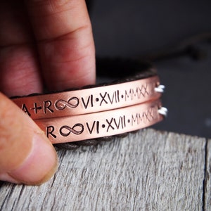 May include: Two copper bracelets with brown leather straps. The bracelets are engraved with the infinity symbol, roman numerals, and the text "A+R 06.17.MMXX".