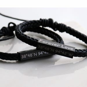 May include: Two black braided bracelets with rectangular metal bars. One bar reads "Forever Love" and the other displays coordinates. The bracelets are on a white surface. The bracelets are a romantic gift.