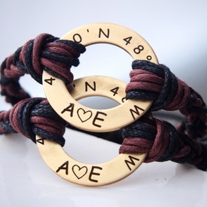 May include: A brown and black braided cord bracelet with two gold metal rings. The rings are engraved with the words "A♥E" and coordinates.