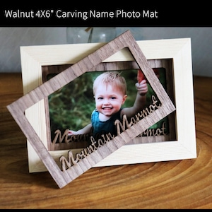 Könnte beinhalten: Ein heller, rechteckiger Bilderrahmen mit einem 10x15 cm großen Foto-Passepartout aus Walnussholz. Das Passepartout liegt schräg über dem Foto und zeigt den lasergeschnittenen Schriftzug "Mountain Marmot" in kursiver Schreibschrift.